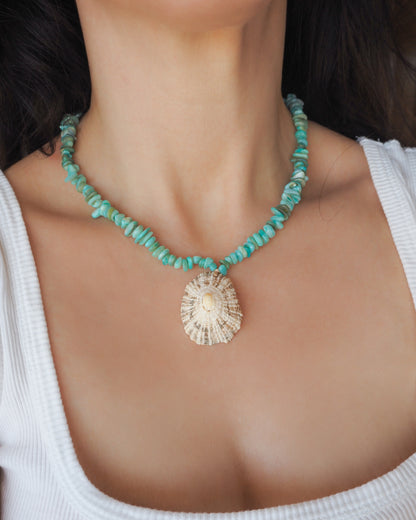 A close-up of a model wearing a turquoise and seashell necklace. The necklace features a strand of small, irregular turquoise beads with a large, textured limpet seashell pendant hanging from a gold-tone clasp. The limpet shell is handpicked from Portugal and has white contrast dark brown stripes