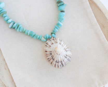 Close up of a beaded necklace with light turquoise-blue stone beads and a large limpet seashell pendant, displayed on a cream fabric surface. The limpet shell is handpicked from Portugal and has white contrast dark brown stripes