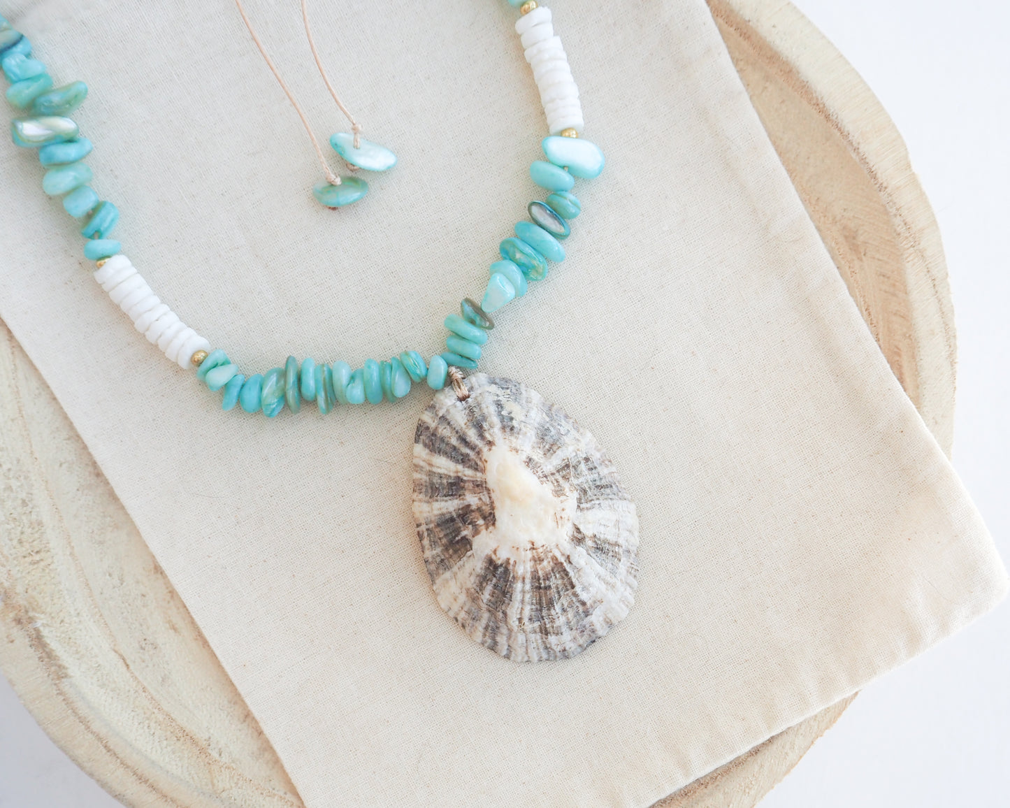 A handmade beaded necklace with turquoise and white beads and a large seashell pendant, displayed on a beige fabric cloth over a wooden surface. The limpet shell is handpicked from Portugal and has white contrast dark brown stripes