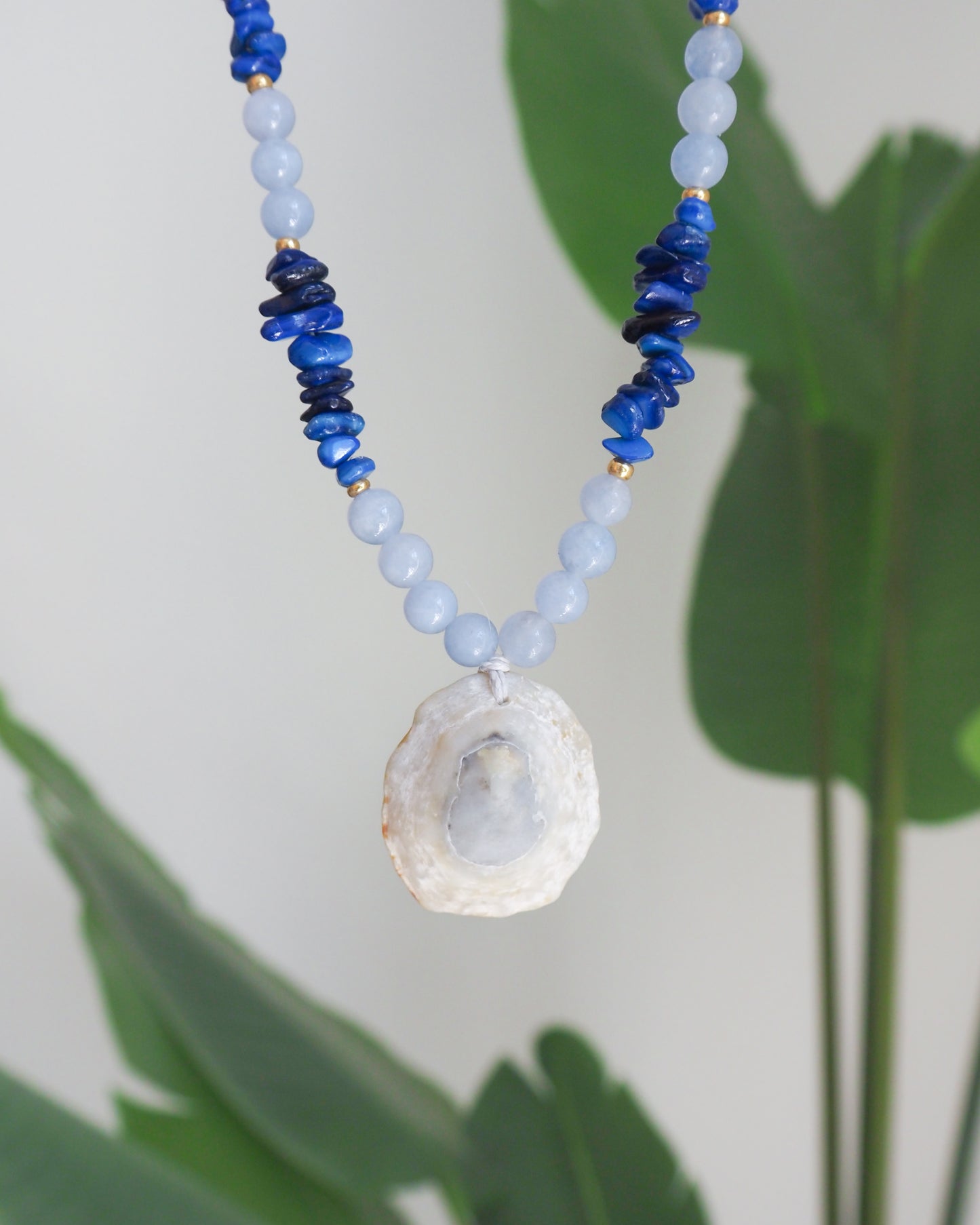 A beaded necklace featuring light blue and dark blue beads, accented with small gold spacers and a natural white clam shell pendant, shown against a soft-focus background of green leaves. A limpet shell necklace with dark blue stone beads and cool blue aquamarine beads. The limpet shell is handpicked from Portugal and has soft grey color with light blueish 