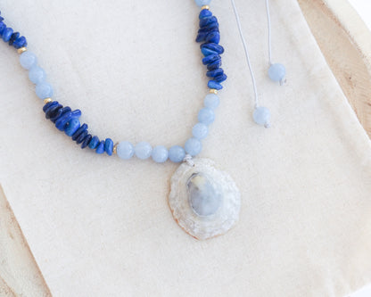 A beaded necklace with light blue and deep blue beads, featuring a large seashell pendant containing a pale blue stone, displayed on a beige fabric background. A limpet shell necklace with dark blue stone beads and cool blue aquamarine beads. The limpet shell is handpicked from Portugal and has soft grey color with light blueish 