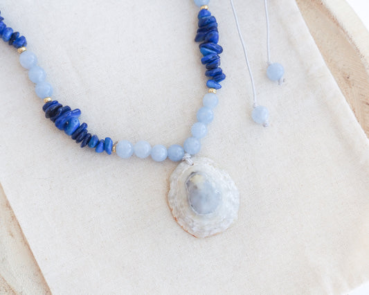A beaded necklace with light blue and deep blue beads, featuring a large seashell pendant containing a pale blue stone, displayed on a beige fabric background. A limpet shell necklace with dark blue stone beads and cool blue aquamarine beads. The limpet shell is handpicked from Portugal and has soft grey color with light blueish 
