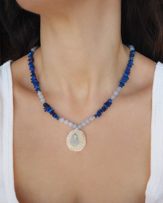 Model wearing a limpet shell necklace with dark blue stone beads and cool blue aquamarine beads. The limpet shell is handpicked from Portugal and has soft grey color with light blueish center.
