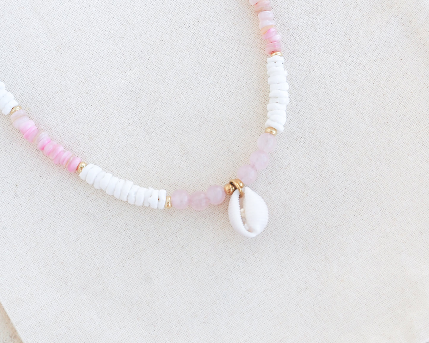 Necklace with pink and white beads on a light background. Close up of a cowrie necklace with soft pink stone beads, rose quartz beads and white shell beads. The center piece of this necklace is a Northern Cowrie Shell, handpicked from the beach of Portugal. The shell is white and about 1 cm. 
