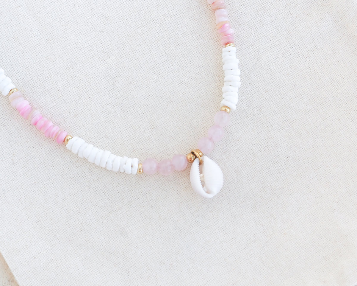 Necklace with pink and white beads on a light background. Close up of a cowrie necklace with soft pink stone beads, rose quartz beads and white shell beads. The center piece of this necklace is a Northern Cowrie Shell, handpicked from the beach of Portugal. The shell is white and about 1 cm. 