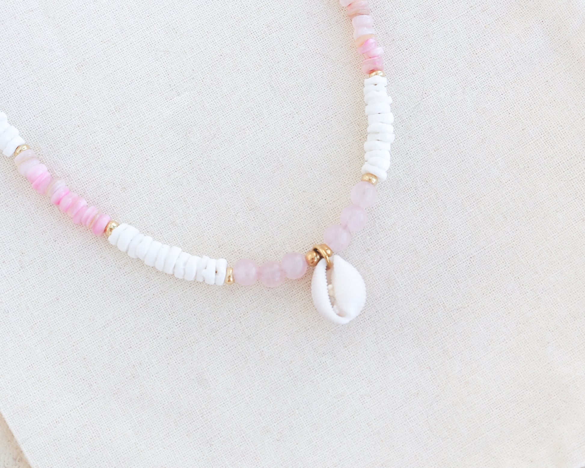 Necklace with pink and white beads on a light background. Close up of a cowrie necklace with soft pink stone beads, rose quartz beads and white shell beads. The center piece of this necklace is a Northern Cowrie Shell, handpicked from the beach of Portugal. The shell is white and about 1 cm. 