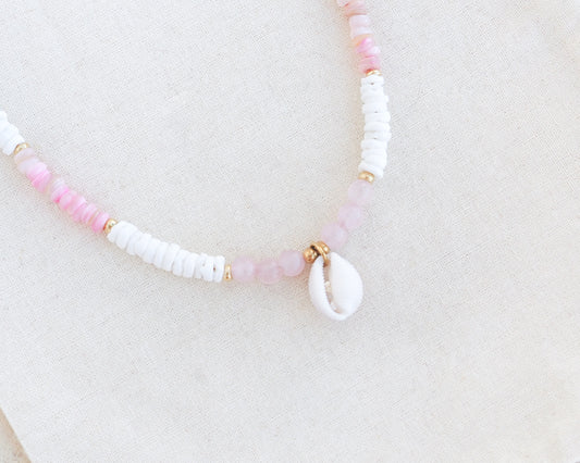 Necklace with pink and white beads on a light background. Close up of a cowrie necklace with soft pink stone beads, rose quartz beads and white shell beads. The center piece of this necklace is a Northern Cowrie Shell, handpicked from the beach of Portugal. The shell is white and about 1 cm. 