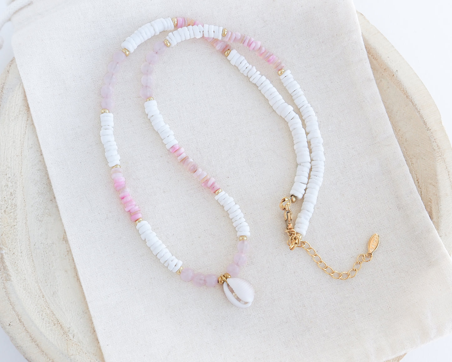 A delicate beaded necklace featuring white textured beads, light pink beads, and a white cowrie shell pendant, accented with small gold spacer beads and a gold chain extender, displayed on a cream fabric cloth.