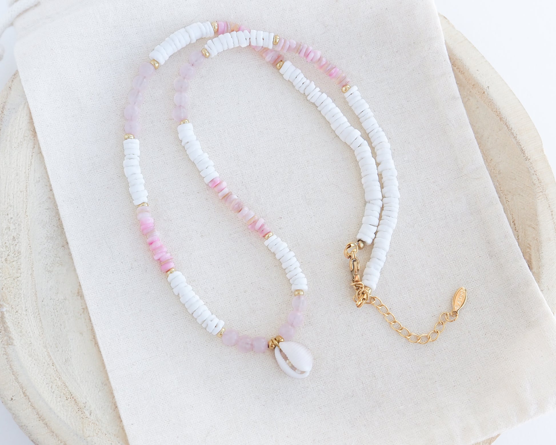 A delicate beaded necklace featuring white textured beads, light pink beads, and a white cowrie shell pendant, accented with small gold spacer beads and a gold chain extender, displayed on a cream fabric cloth.