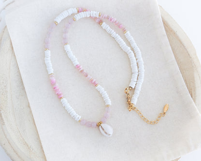A delicate beaded necklace featuring white textured beads, light pink beads, and a white cowrie shell pendant, accented with small gold spacer beads and a gold chain extender, displayed on a cream fabric cloth.