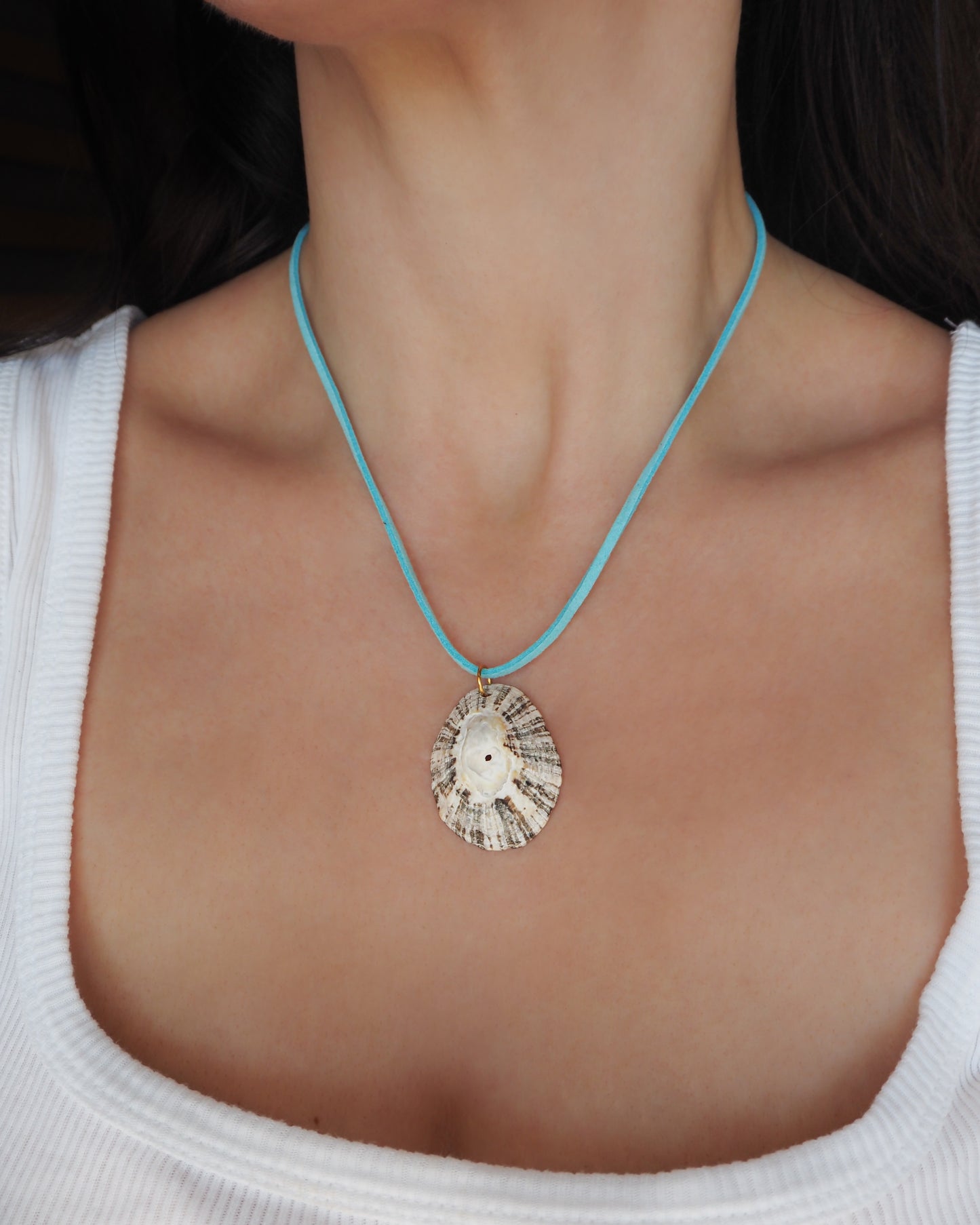 Model wearing a seashell pendant necklace with a light blue cord and white beads, displayed on a textured cream fabric background. The shell is a limpet shell from Portugal and is about 5 cm. The color of the shell is dark and white and it has stripes. 