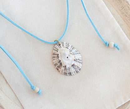 A seashell pendant necklace with a light blue cord and white beads, displayed on a textured cream fabric background. The shell is a limpet shell from Portugal and is about 5 cm. The color of the shell is dark and white and it has stripes. 