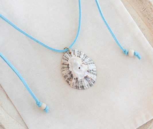 A seashell pendant necklace with a light blue cord and white beads, displayed on a textured cream fabric background. The shell is a limpet shell from Portugal and is about 5 cm. The color of the shell is dark and white and it has stripes. 