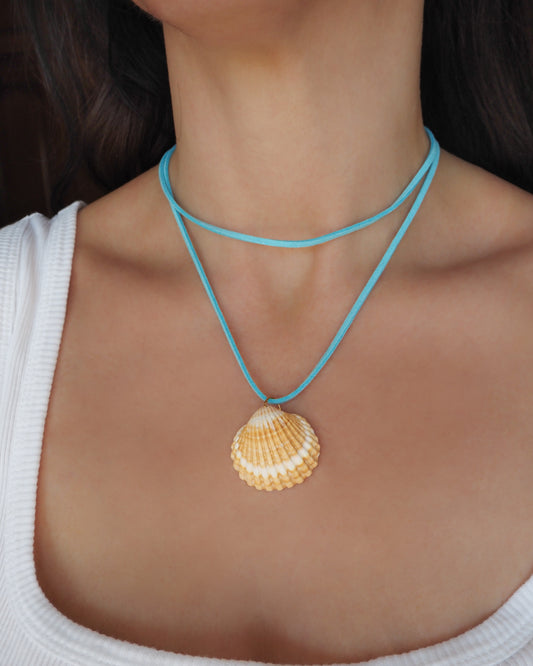 A close-up of a model wearing a double-strand turquoise blue cord necklace with a natural beige and white seashell pendant. The shell is a mediterranean cockle shell from Portugal and is about 5 cm. The color of the shell is yellow white and it has vertical ridges. 