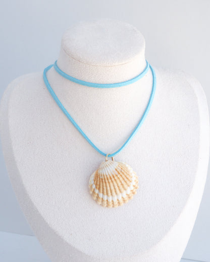 A light blue cord necklace with a natural scallop shell pendant, displayed on a white textured jewelry bust. The shell is a mediterranean cockle shell from Portugal and is about 5 cm. The color of the shell is yellow white and it has vertical ridges. 