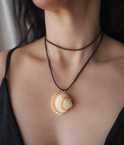 A close-up of a person wearing a brown cord necklace with a realistic seashell pendant, styled over a black camisole top. The shell is a mediterranean cockle shell from Portugal. The shell has yellow white colors and ridges. 