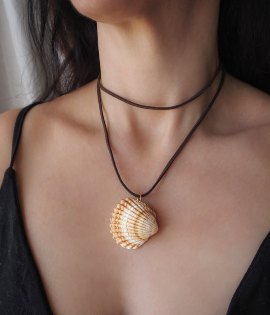 A close-up of a person wearing a brown cord necklace with a realistic seashell pendant, styled over a black camisole top. The shell is a mediterranean cockle shell from Portugal. The shell has yellow white colors and ridges. 