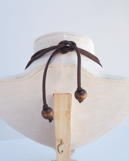 A dark brown cord choker necklace with two wooden bead accents, displayed on a cream-colored fabric jewelry bust. The shell is a mediterranean cockle shell from Portugal. The shell has yellow white colors and ridges. 