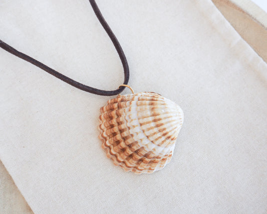 A natural clam shell pendant necklace with a dark brown cord, displayed on a textured beige fabric background. The shell is a mediterranean cockle shell from Portugal. The shell has yellow white colors and ridges. 