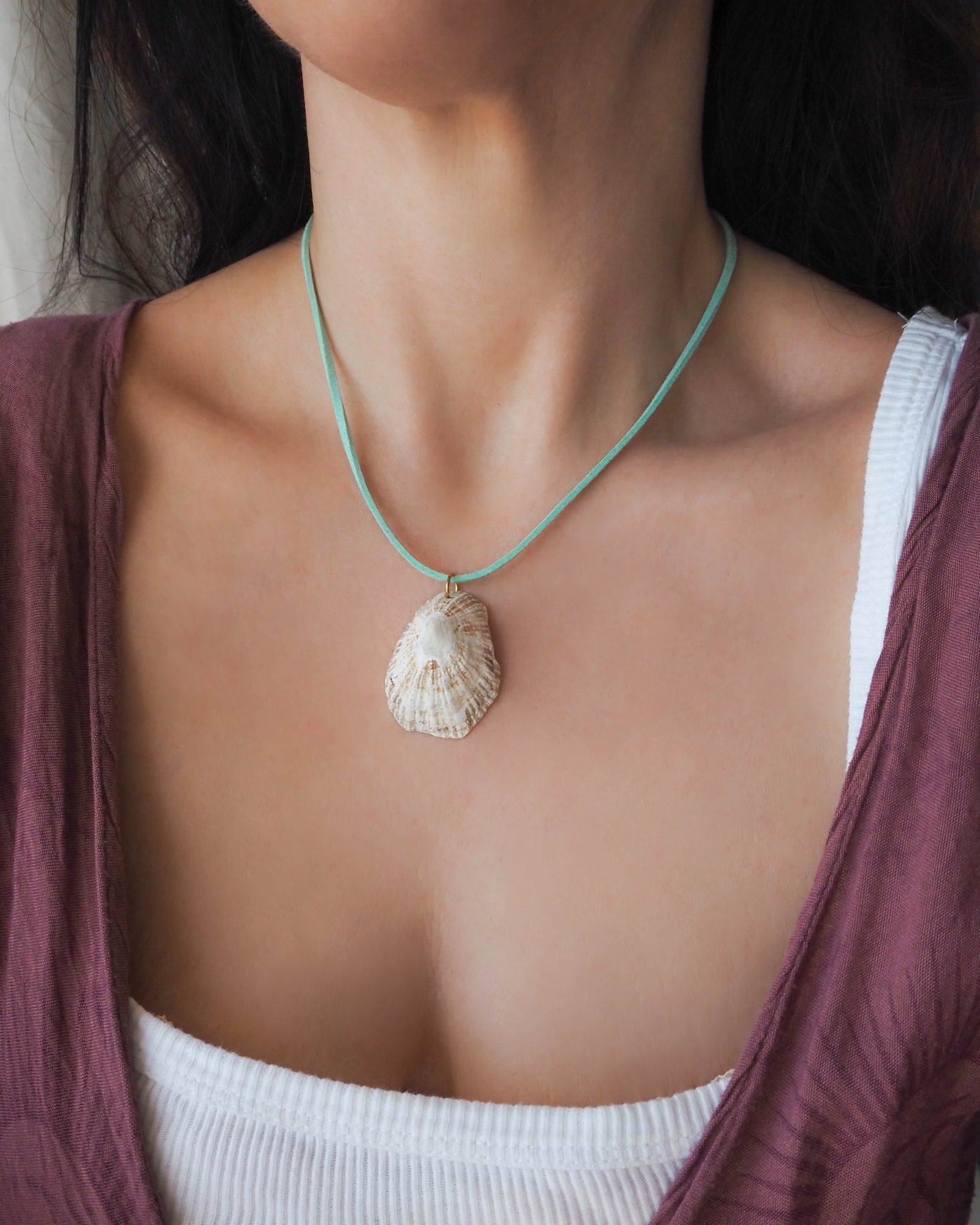 A close-up of a person wearing a natural seashell pendant necklace on a turquoise cord, styled over a white ribbed tank top and a mauve cardigan.