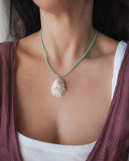 A close-up of a person wearing a natural seashell pendant necklace on a turquoise cord, styled over a white ribbed tank top and a mauve cardigan.