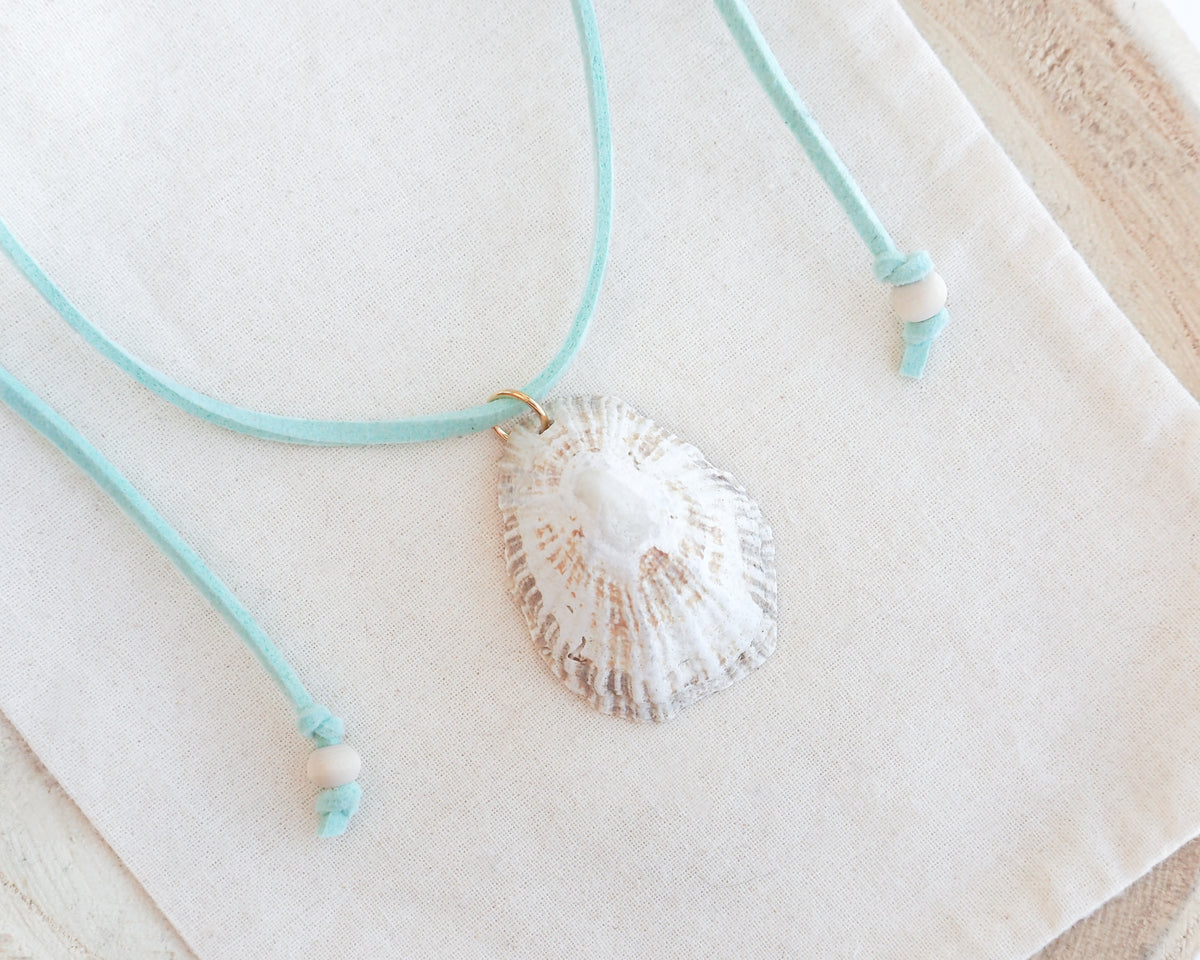 A limpet seashell pendant necklace with a mint turquoise cord and small white beads, displayed on a textured cream fabric background.