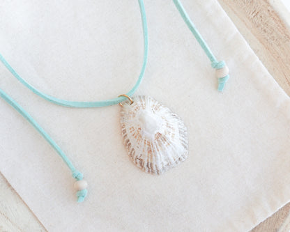 A limpet seashell pendant necklace with a mint turquoise cord and small white beads, displayed on a textured cream fabric background.