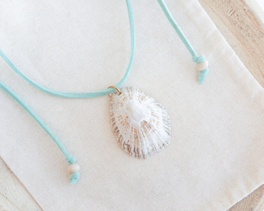 A limpet seashell pendant necklace with a mint turquoise cord and small white beads, displayed on a textured cream fabric background.