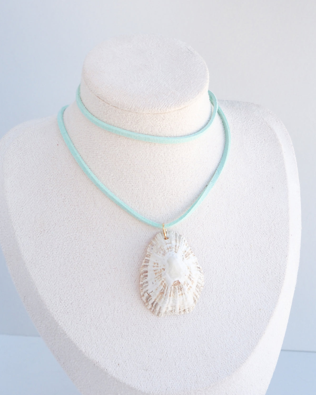 A necklace displayed on a white jewelry bust, featuring a turquoise cord with a large white seashell pendant and a thin mint turquoise wrap around the neck area.