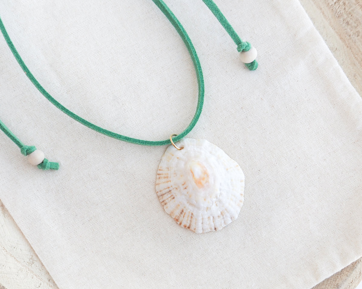 A seashell pendant necklace with a green cord and white beads, displayed on a textured cream fabric background. The shell is a limpet shell from Portugal and is about 5 cm. The color of the shell is off white and it has an orange center.
