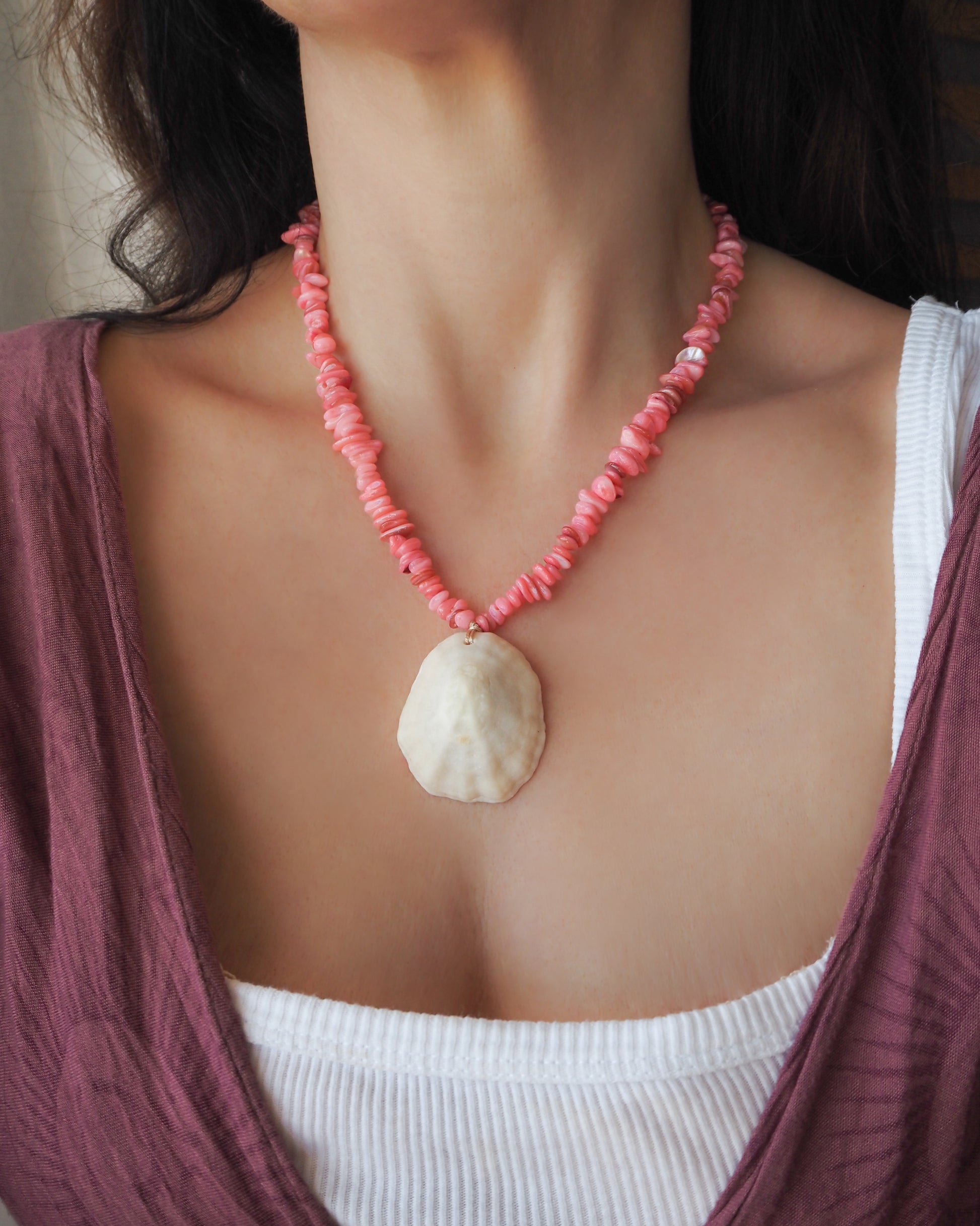 A close-up of a person wearing a necklace with pink coral-like beads and a large ivory white-colored shell pendant, layered over a white ribbed top and a mauve cardigan. The shell is a limpet shell handpicked from the beach of Portugal and is 5 cm.