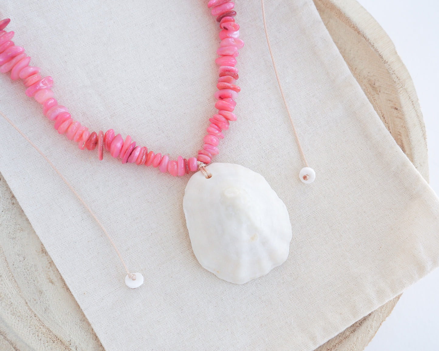 A pink beaded necklace with a large white clam shell pendant, laid flat on a beige linen cloth over a round wooden surface. The shell is a limpet shell handpicked from the beach of Portugal and is 5 cm.