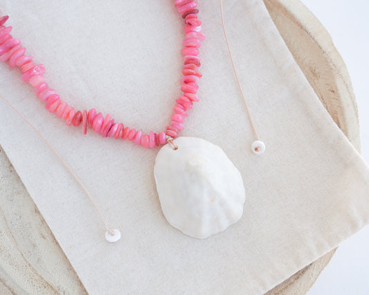 A pink beaded necklace with a large white clam shell pendant, laid flat on a beige linen cloth over a round wooden surface. The shell is a limpet shell handpicked from the beach of Portugal and is 5 cm.