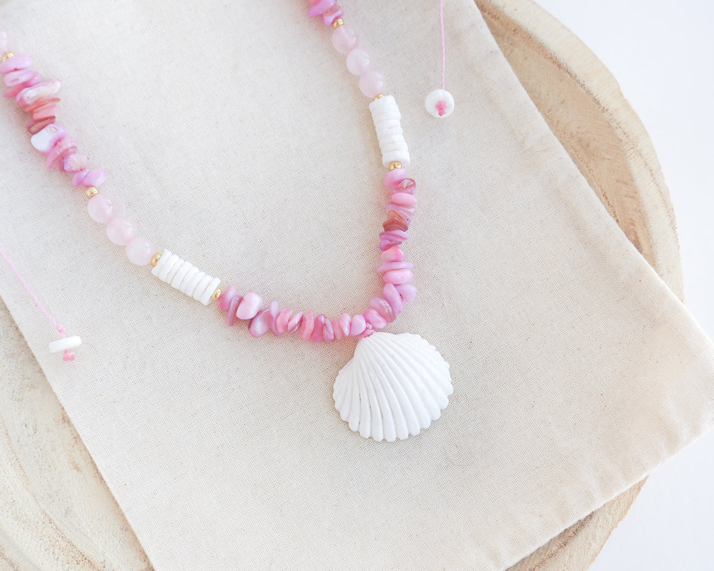a pink and white beaded necklace with a large seashell pendant, displayed on a beige fabric surface. A necklace with pink and white beads and a large cone shell pendant on a light background. A necklace with a mediterranean cockle shell paired with rose quartz gemstone beads, soft pink stones and white shell beads. The shell is a mediterranean cockle shell from Portugal and is about 3 cm. The color of the shell is white.