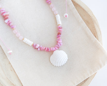 a pink and white beaded necklace with a large seashell pendant, displayed on a beige fabric surface. A necklace with pink and white beads and a large cone shell pendant on a light background. A necklace with a mediterranean cockle shell paired with rose quartz gemstone beads, soft pink stones and white shell beads. The shell is a mediterranean cockle shell from Portugal and is about 3 cm. The color of the shell is white.