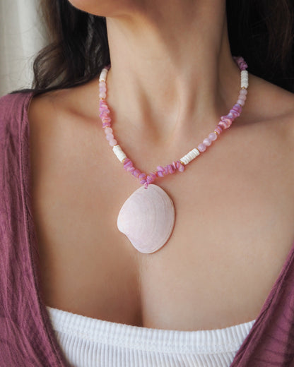 Model wearing a pink and white beaded necklace with a large seashell pendant. A necklace with pink and white beads and a large cone shell pendant on a light background. A necklace with a clam shell paired with rose quartz gemstone beads, soft pink stones and white shell beads. The shell is a smooth clam shell from Portugal and is about 6 cm. The color of the shell is soft pink and is very unique for these shells.