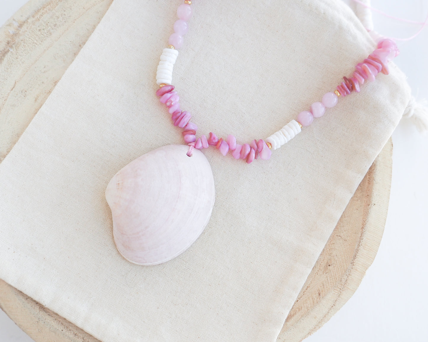 a pink and white beaded necklace with a large seashell pendant, displayed on a beige fabric surface. A necklace with pink and white beads and a large cone shell pendant on a light background. A necklace with a clam shell paired with rose quartz gemstone beads, soft pink stones and white shell beads. The shell is a smooth clam shell from Portugal and is about 6 cm. The color of the shell is soft pink and is very unique for these shells.
