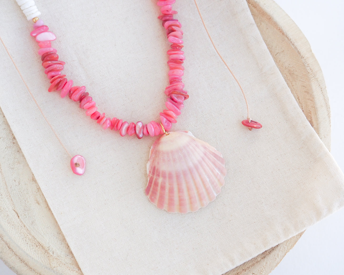 Close up of a necklace with pink and white beads and a shell pendant on light cotton displayed. A necklace with a big pink scallop shell paired with rose quartz gemstone beads, coral pink stones and white shell beads. The shell is a pilgrim's scallop shell handpicked from Portugal and is about 5 cm. The color of the shell is soft pink.