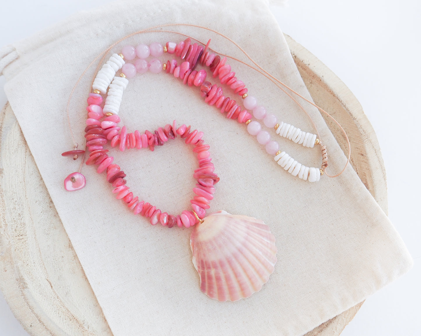 A pink and white beaded necklace with a large scallop shell pendant, displayed on a cream fabric surface over a round wooden tray. Close up of a necklace with pink and white beads and a shell pendant on light cotton displayed. A necklace with a big pink scallop shell paired with rose quartz gemstone beads, coral pink stones and white shell beads. The shell is a pilgrim's scallop shell handpicked from Portugal and is about 5 cm. The color of the shell is soft pink.
