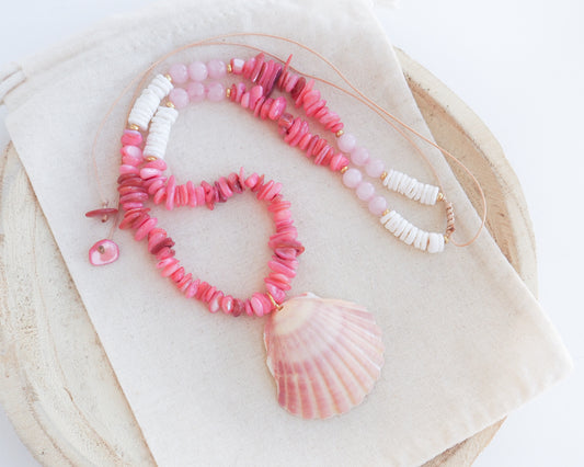 A pink and white beaded necklace with a large scallop shell pendant, displayed on a cream fabric surface over a round wooden tray. Close up of a necklace with pink and white beads and a shell pendant on light cotton displayed. A necklace with a big pink scallop shell paired with rose quartz gemstone beads, coral pink stones and white shell beads. The shell is a pilgrim's scallop shell handpicked from Portugal and is about 5 cm. The color of the shell is soft pink.