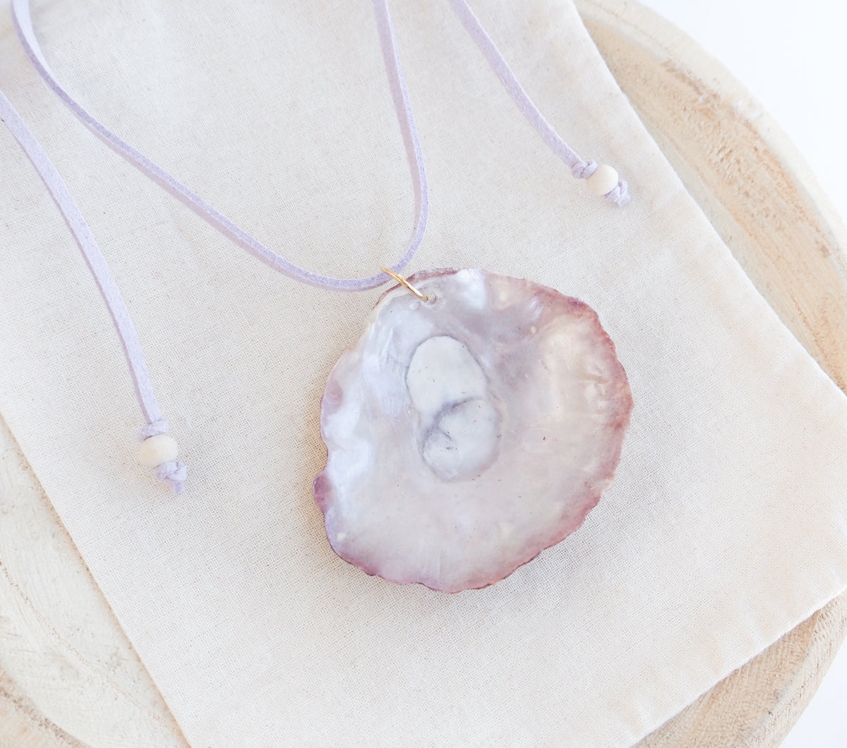 A close-up of a light pink cord choker necklace with a large, pearly shell pendant. It's made with a jingle shell handpicked from the beaches of Portugal. The necklace is made of suede. 