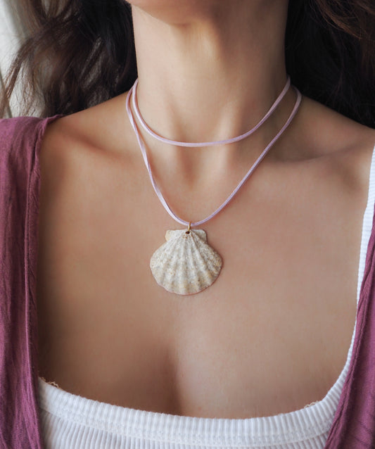 A close-up of a model wearing a double-layered necklace featuring a natural beige scallop seashell pendant on a light pink cord, styled over a white ribbed tank top and a mauve cardigan.