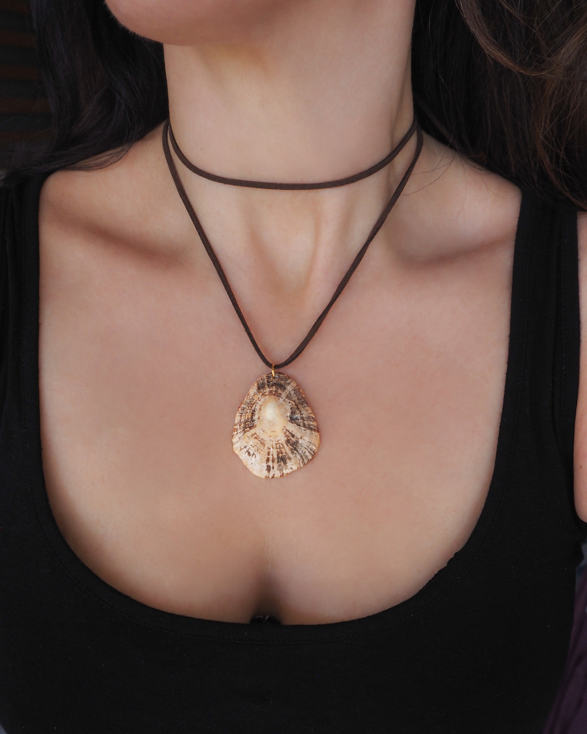 A close-up of a person wearing a dark brown double-layered cord necklace with a natural beige and brown seashell pendant. A seashell pendant necklace with a brown suede cord, displayed on neck. The shell is a limpet shell from Portugal and is about 5 cm. The color of the shell is brown beige and it has ridges.