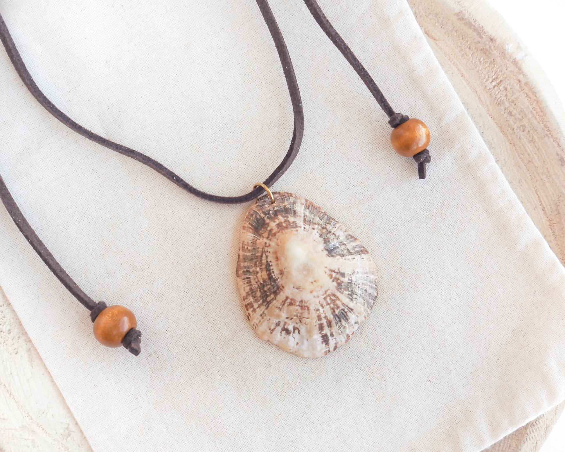 A natural limpet shell pendant necklace on a dark brown cord with wooden beads, displayed on a textured cream linen cloth. A seashell pendant necklace with a brown suede cord. The shell is a limpet shell from Portugal and is about 5 cm. The color of the shell is brown beige and it has ridges.