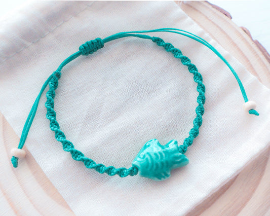 A close-up photo of the Fish Bracelet's intricate ceramic fish charm with shimmering green enamel detailing, Ceramic fish bead, green turquoise braided braccelet