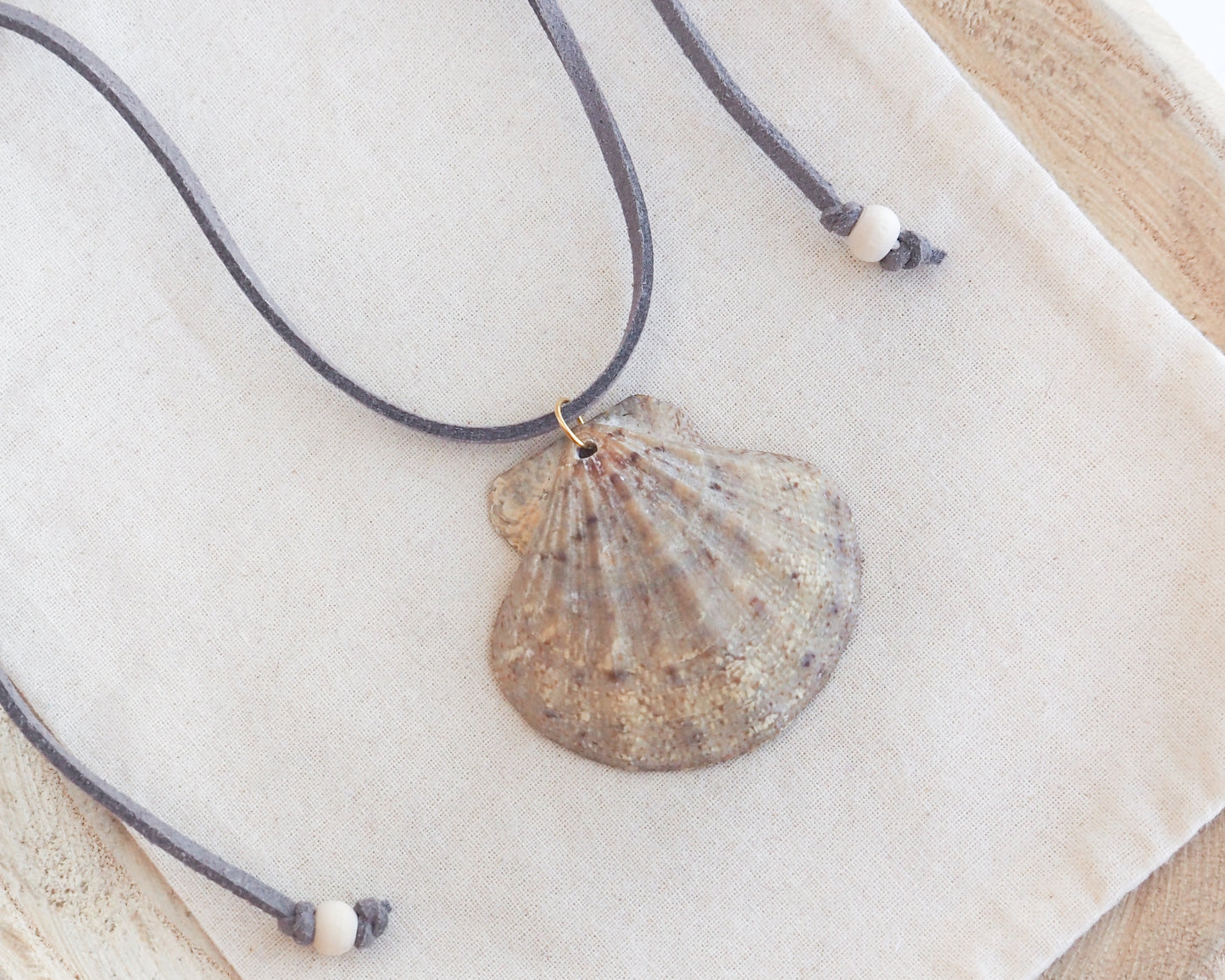 A scallop shell pendant necklace on a gray suede cord with white beads, displayed on a textured cream linen cloth.