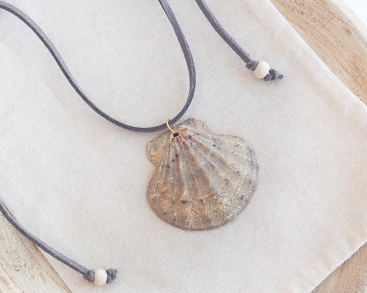 A scallop shell pendant necklace on a gray suede cord with white beads, displayed on a textured cream linen cloth.
