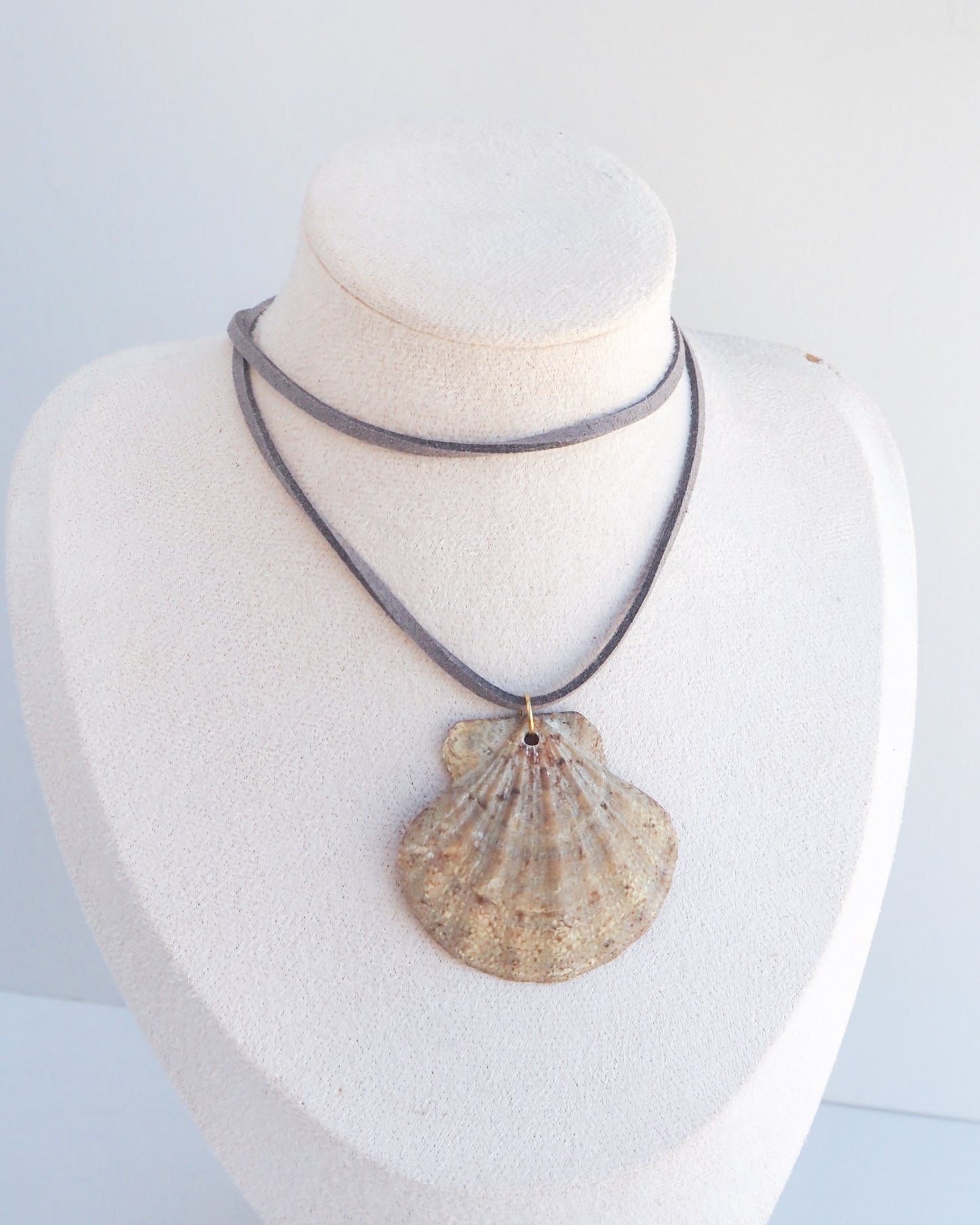 A shell-shaped pendant necklace displayed on a white fabric jewelry bust. The pendant has a natural scallop shell design in beige and brown tones, suspended from a layered gray cord necklace.