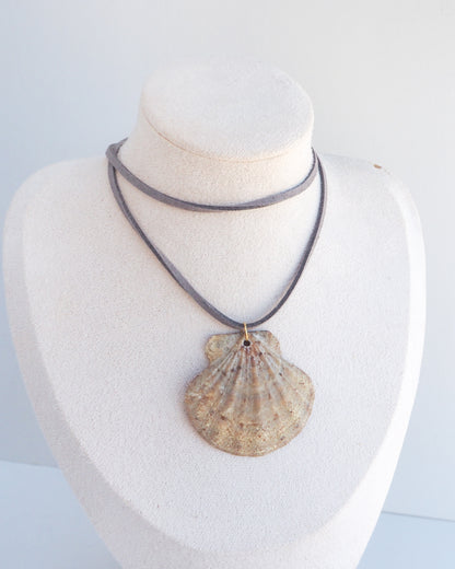 A shell-shaped pendant necklace displayed on a white fabric jewelry bust. The pendant has a natural scallop shell design in beige and brown tones, suspended from a layered gray cord necklace.