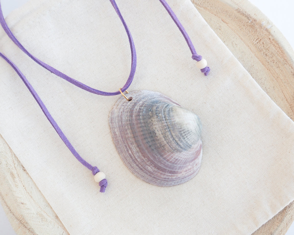 A purple cord necklace with a natural clam shell pendant, displayed on a cream fabric pouch over a light wood surface. The shell is a Violet Bittersweet Clam Shell handpicked from the beaches of Portugal.